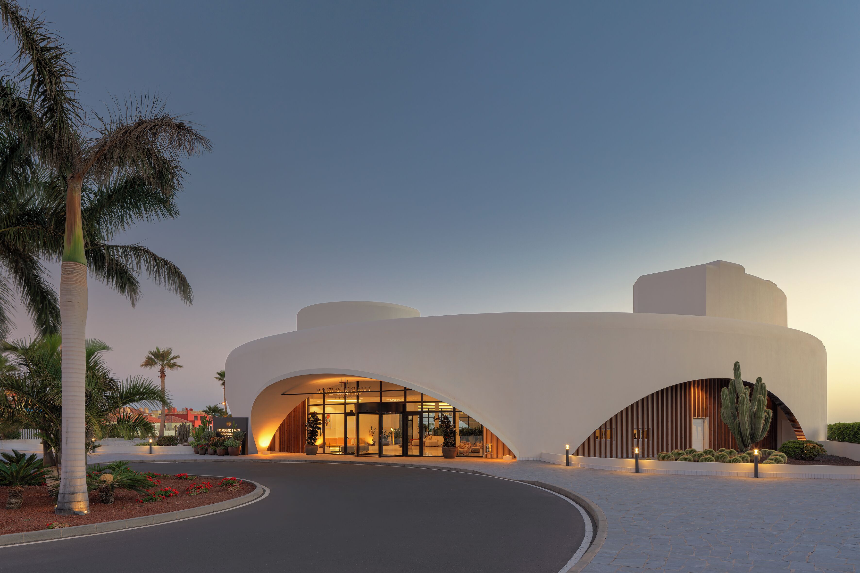 Un hotel moderno con una arquitectura curva distintiva, una entrada bien iluminada y un área ajardinada con cactus y palmeras.