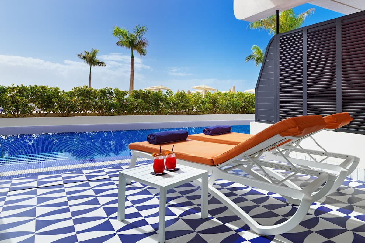 A poolside area with a geometric tile floor, a lounge chair, a table with drinks, and a view of palm trees and a clear sky.