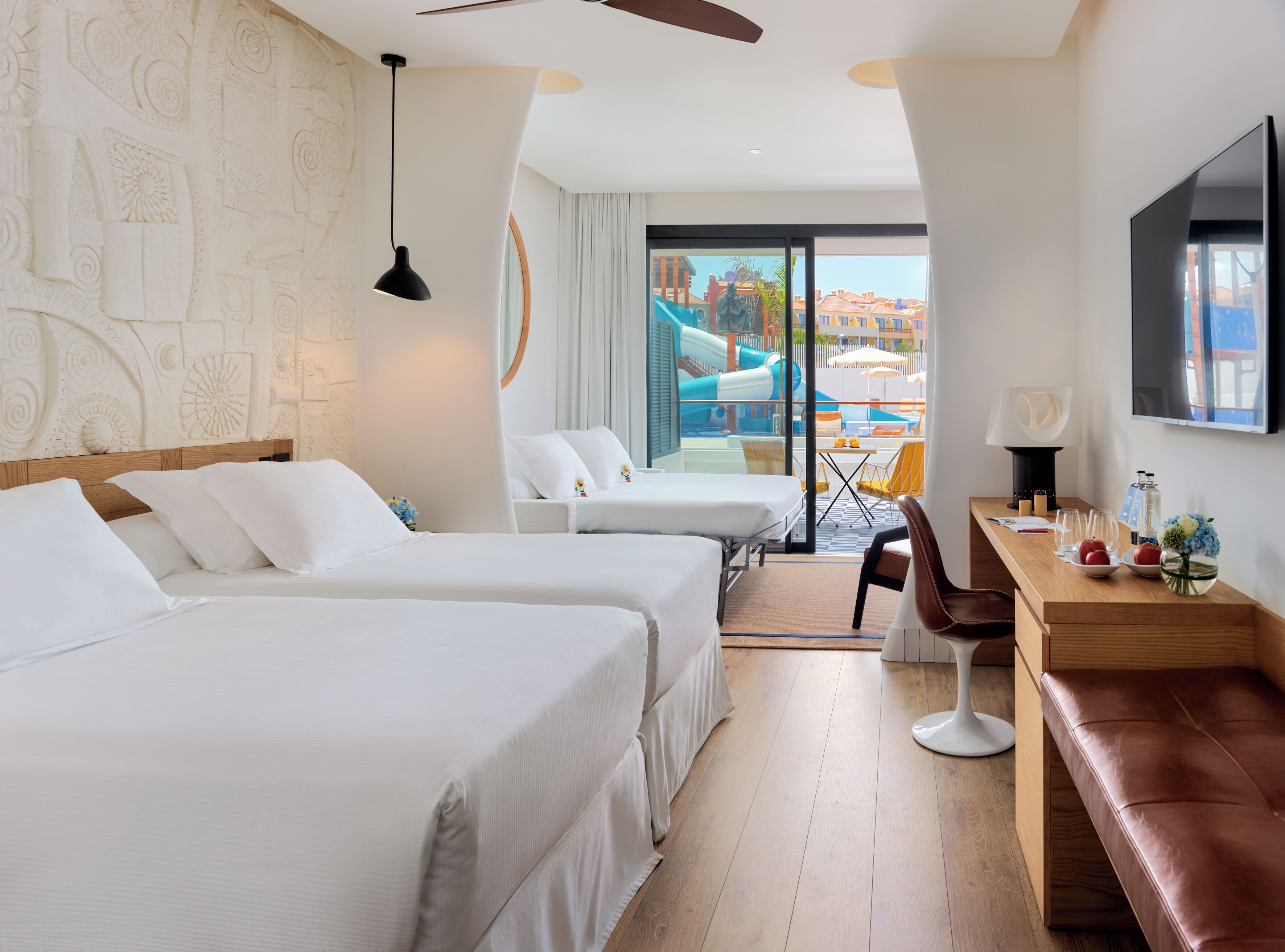 Habitación de hotel con dos camas, un escritorio, una silla, un televisor y vistas a una piscina y edificios.