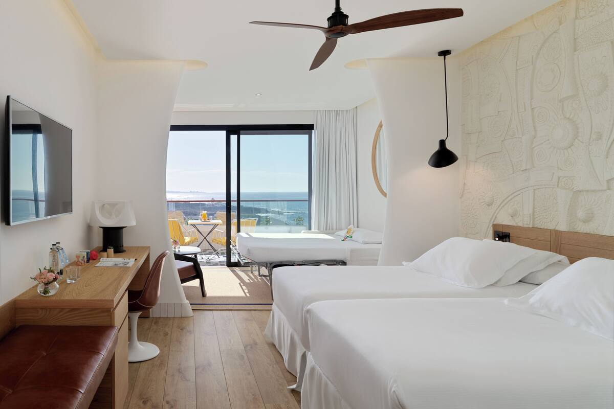 A hotel room with a bed, a desk, a chair, a TV, a ceiling fan, and a view of the ocean.