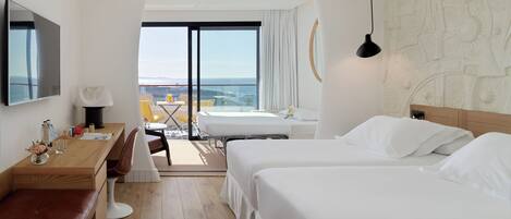 A hotel room with a bed, a desk, a chair, a TV, a ceiling fan, and a view of the ocean.