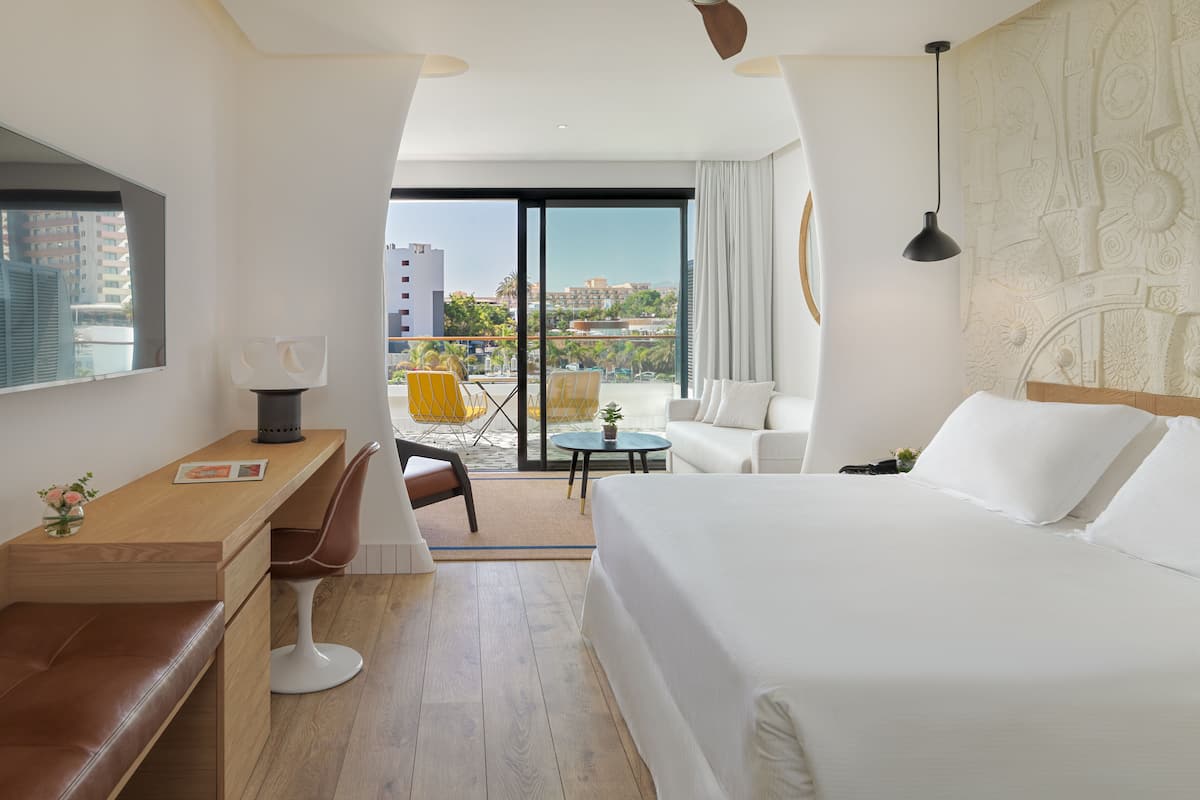 A modern hotel room with a large bed, a desk, a chair, a small table, and a view of the city through large windows.