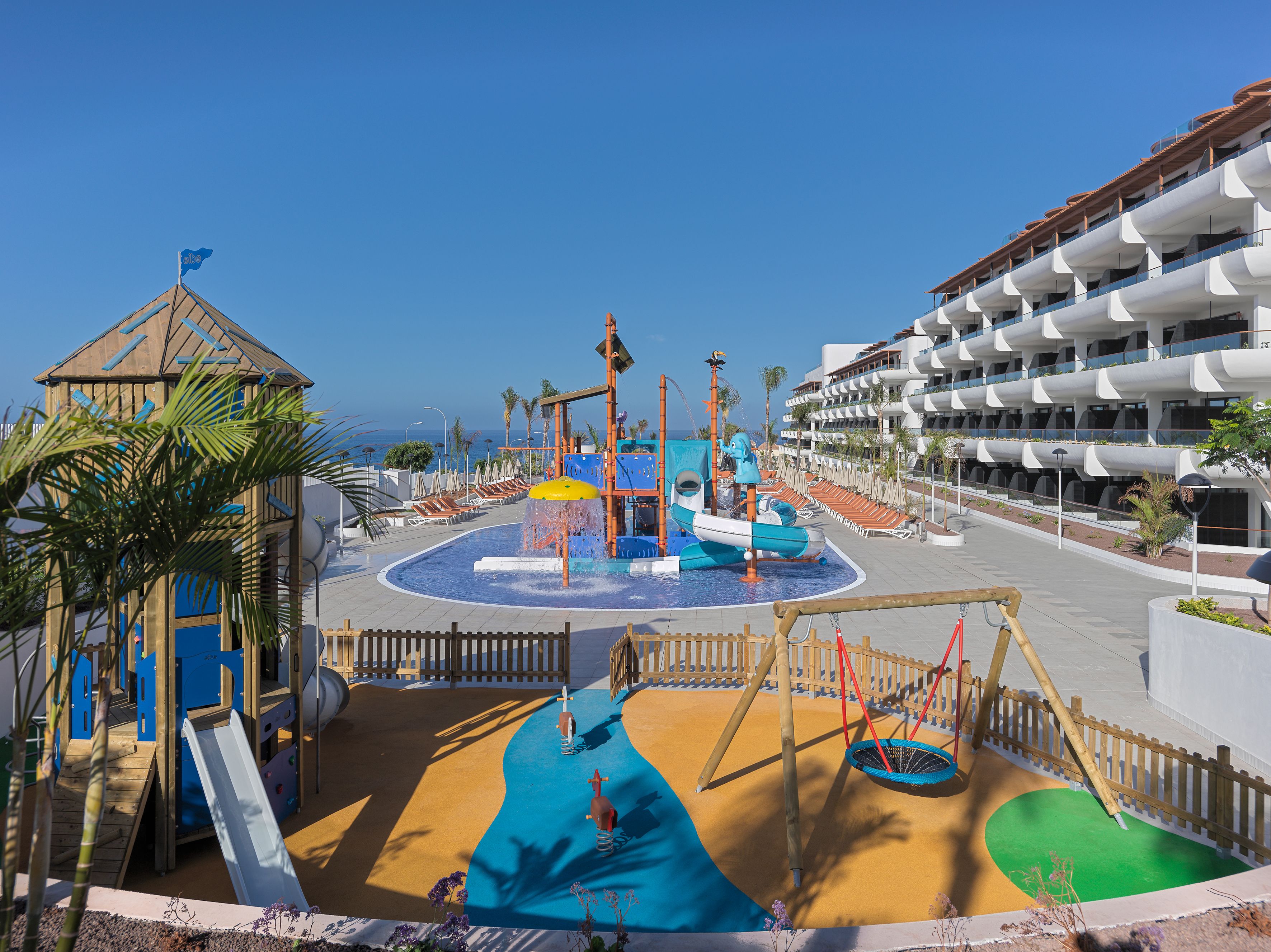 Un espace de jeux pour enfants avec des toboggans et des jeux d’eau, entouré de palmiers et bordé d’un bâtiment doté de balcons.