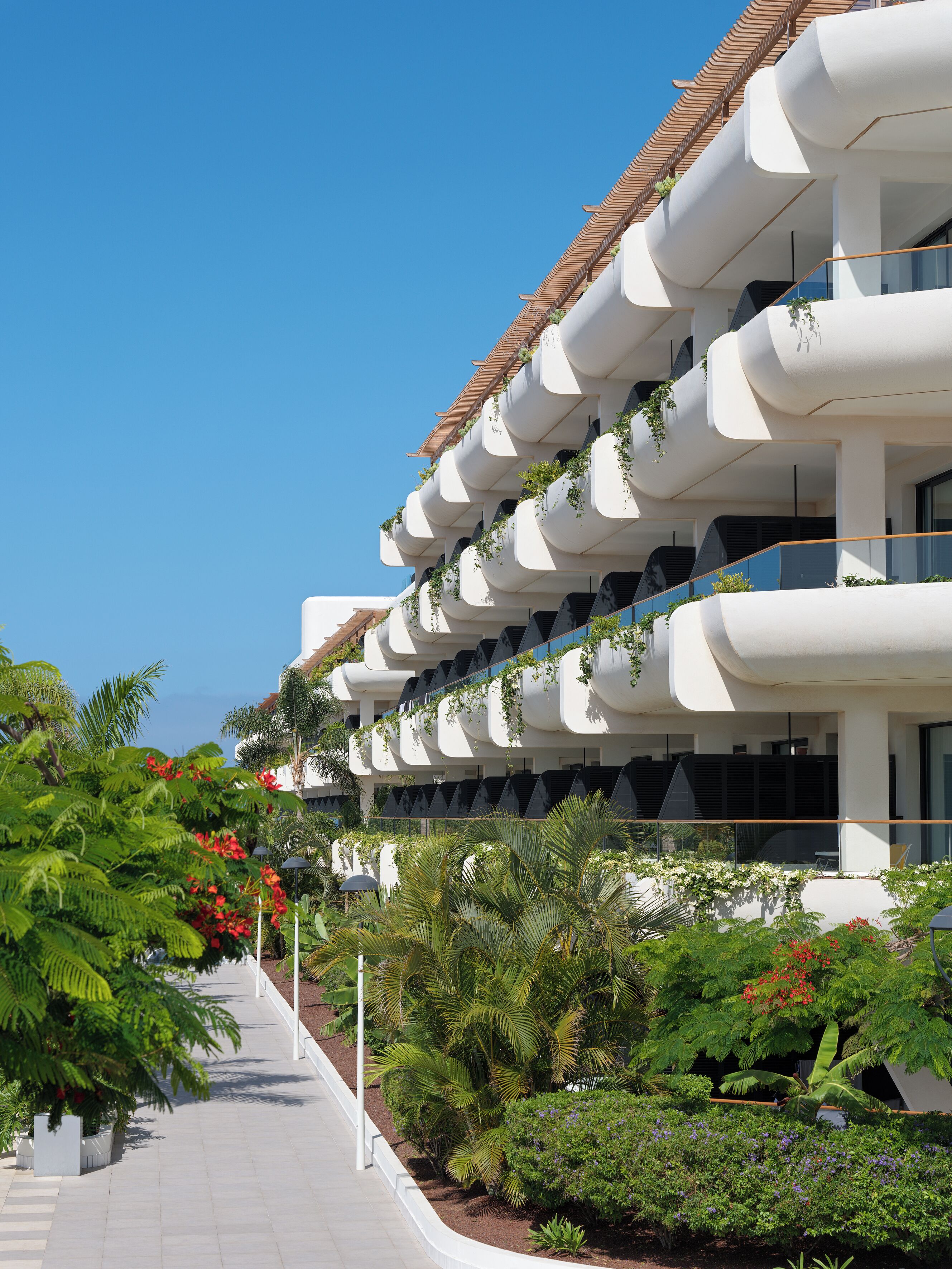 Um edifício moderno com várias varandas, algumas com plantas, e um caminho ladeado por plantas e flores tropicais.