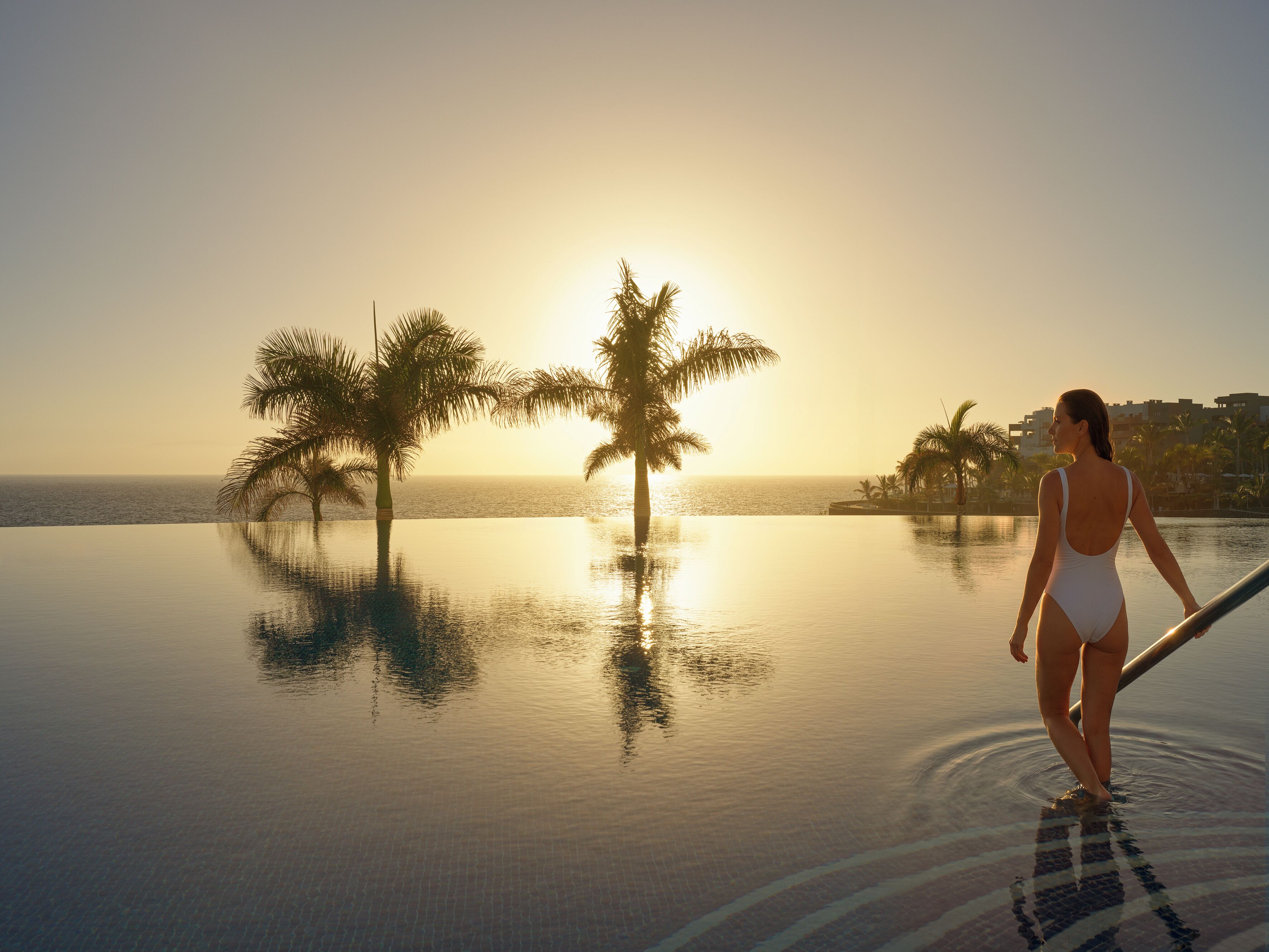 Una mujer en traje de baño de pie junto a una piscina infinita, con palmeras y un atardecer al fondo.