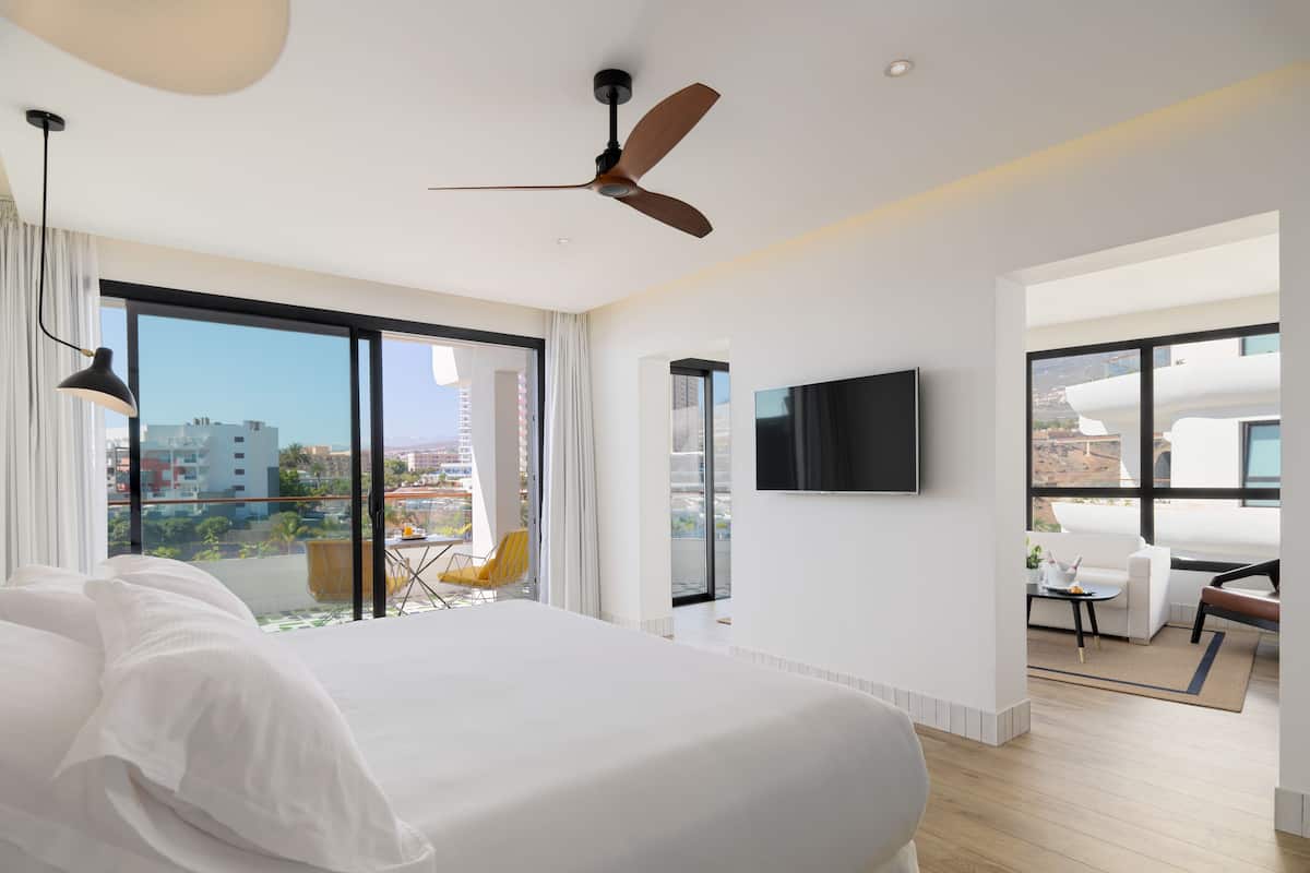A modern hotel room with a large bed, a ceiling fan, a TV mounted on the wall, and a balcony with a view of the city.