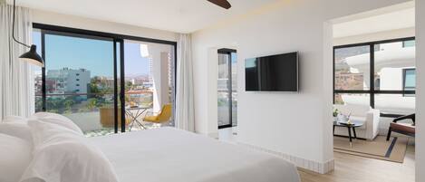 A modern hotel room with a large bed, a ceiling fan, a TV mounted on the wall, and a balcony with a view of the city.