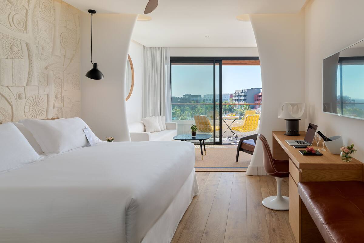 A hotel room with a large bed, a desk with a laptop, a chair, and a view of the city through large windows.