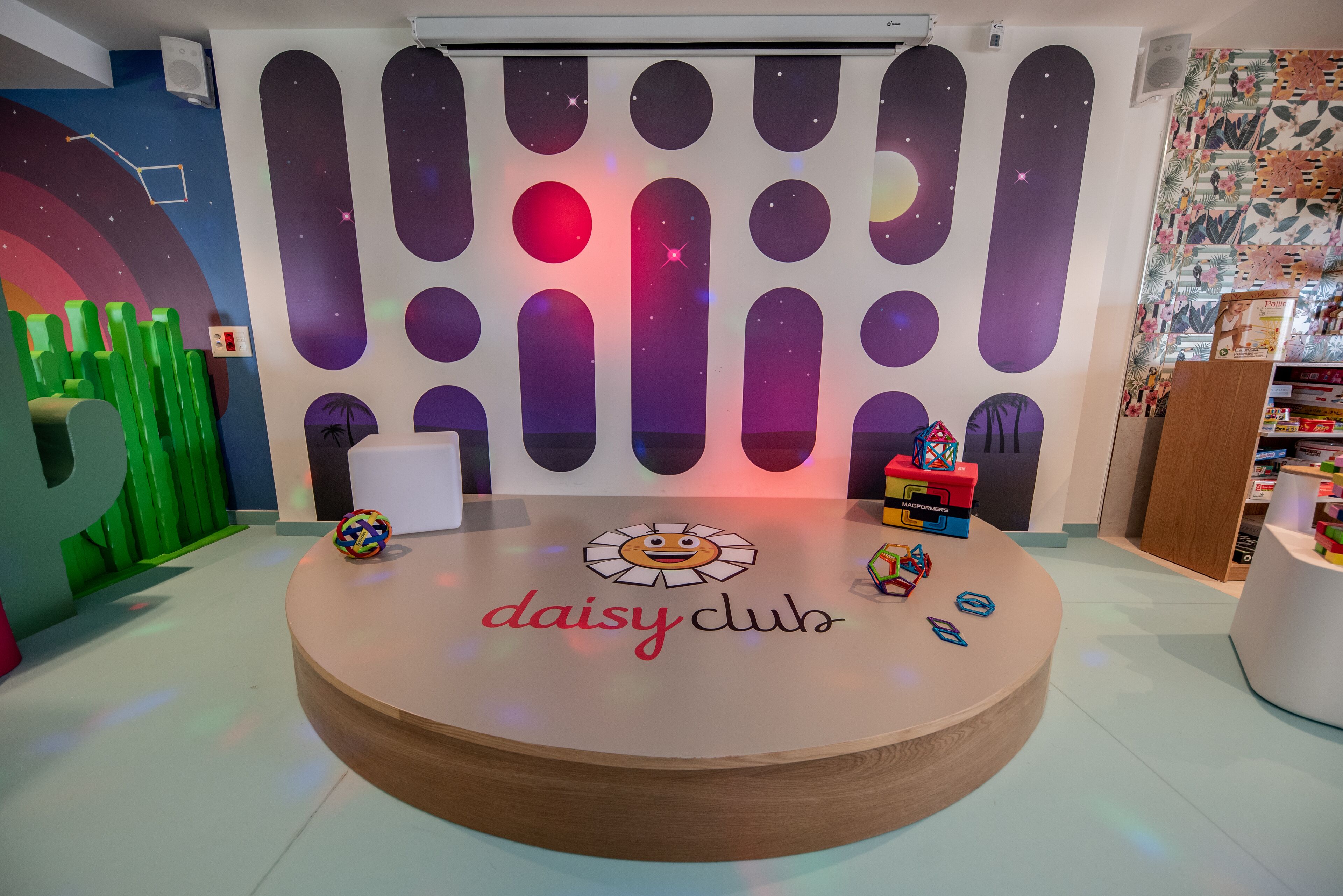 A circular wooden table with the text "daisy club" in the center, surrounded by colorful toys and a wall with a space-themed design.
