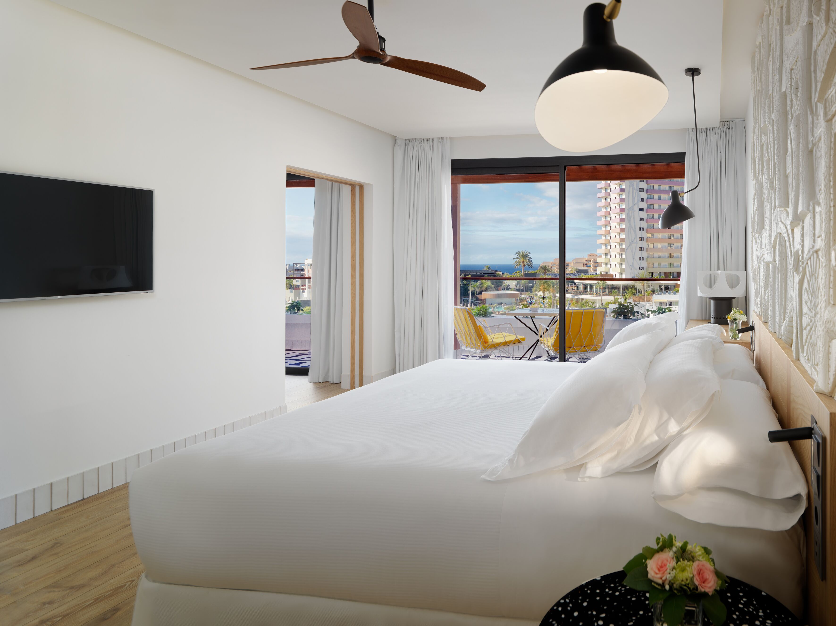 Habitació d'hotel moderna amb un llit gran, un ventilador de sostre, una televisió de pantalla plana i un balcó amb cadires grogues.