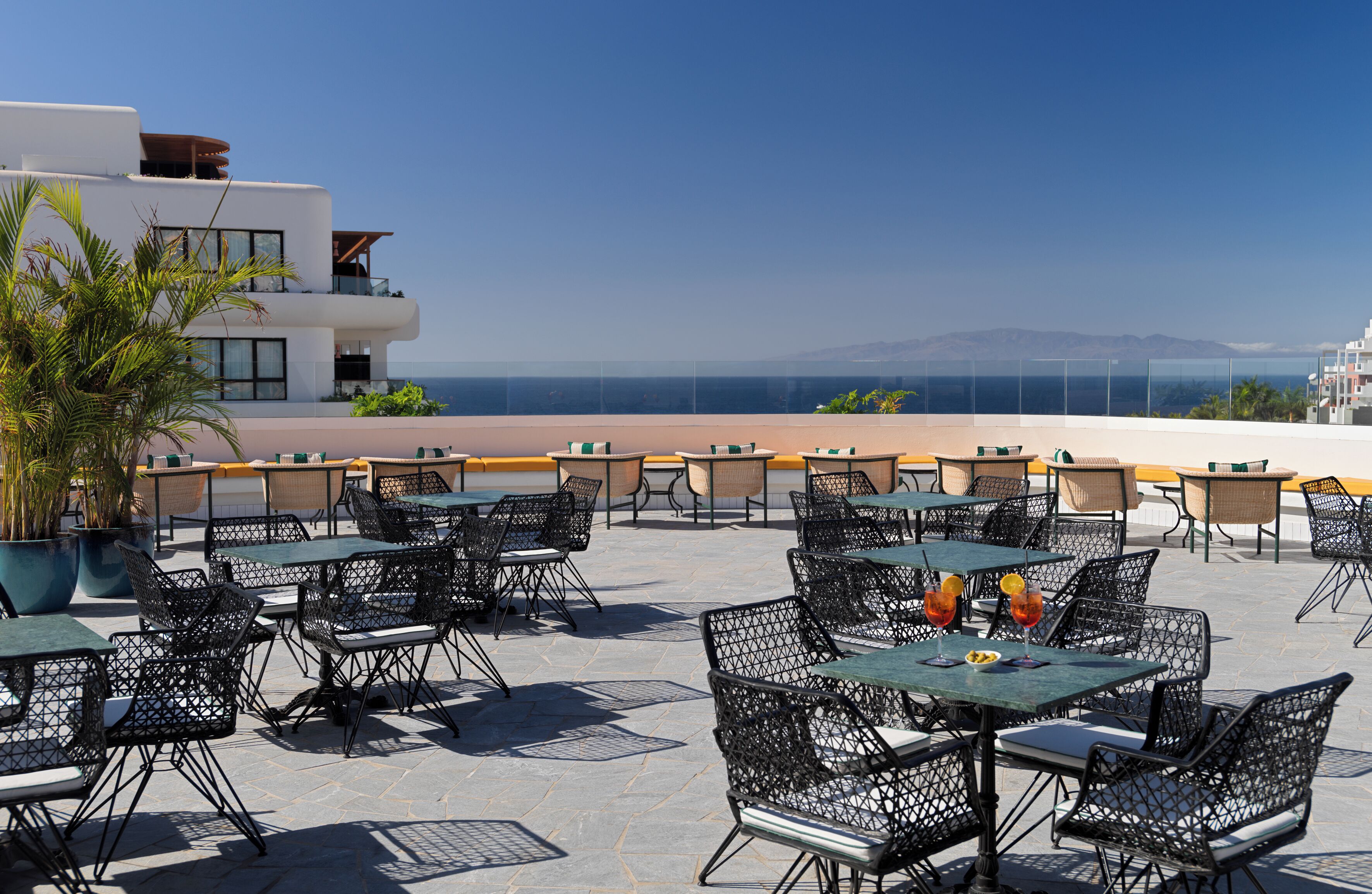 An outdoor seating area with tables and chairs, overlooking a body of water and a mountainous landscape.