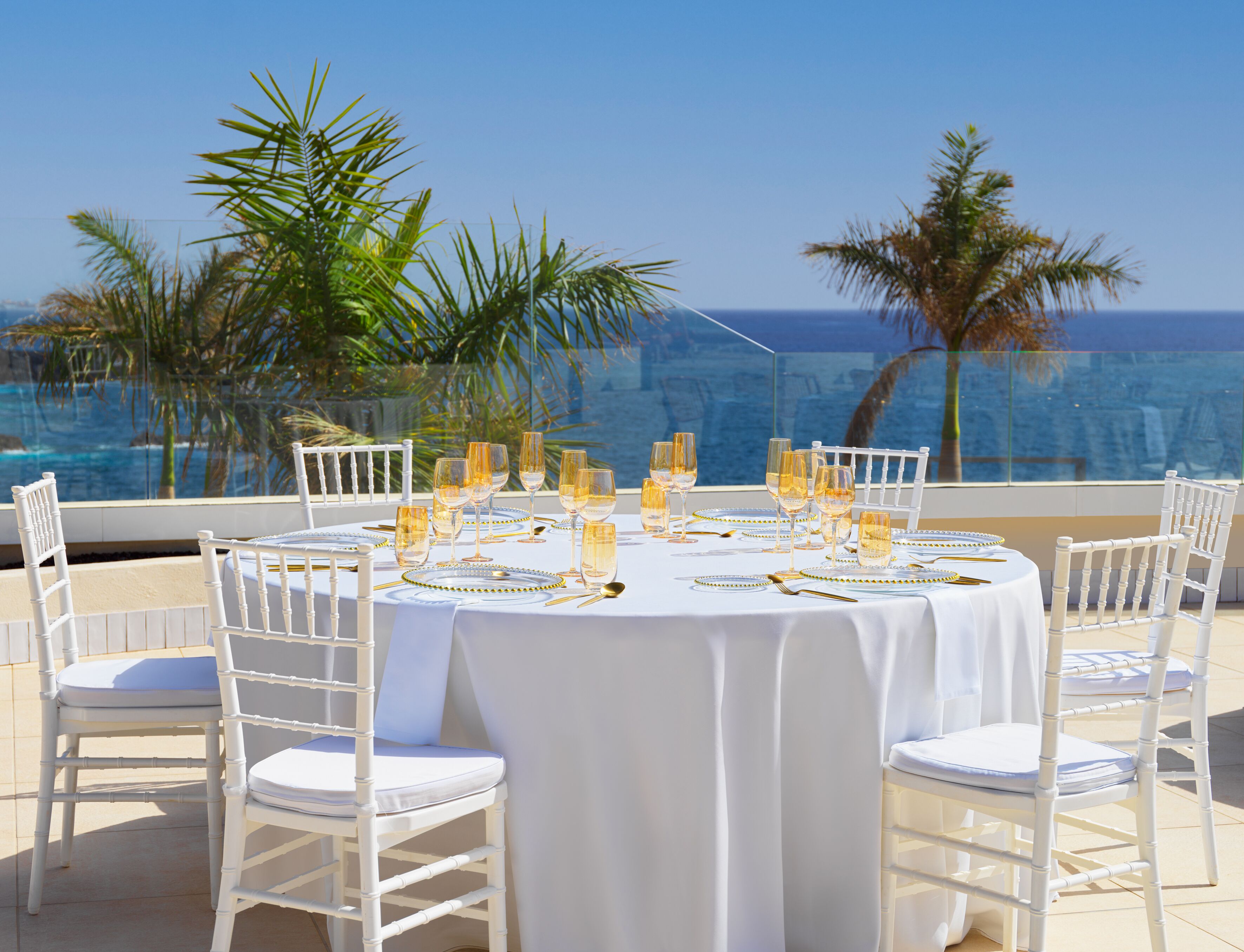 A round table set for a meal with white chairs, overlooking a body of water with palm trees.