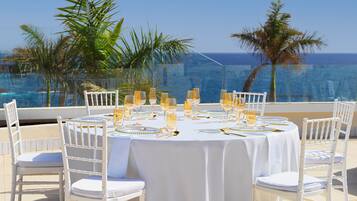 A round table set for a meal with white chairs, overlooking a body of water with palm trees.