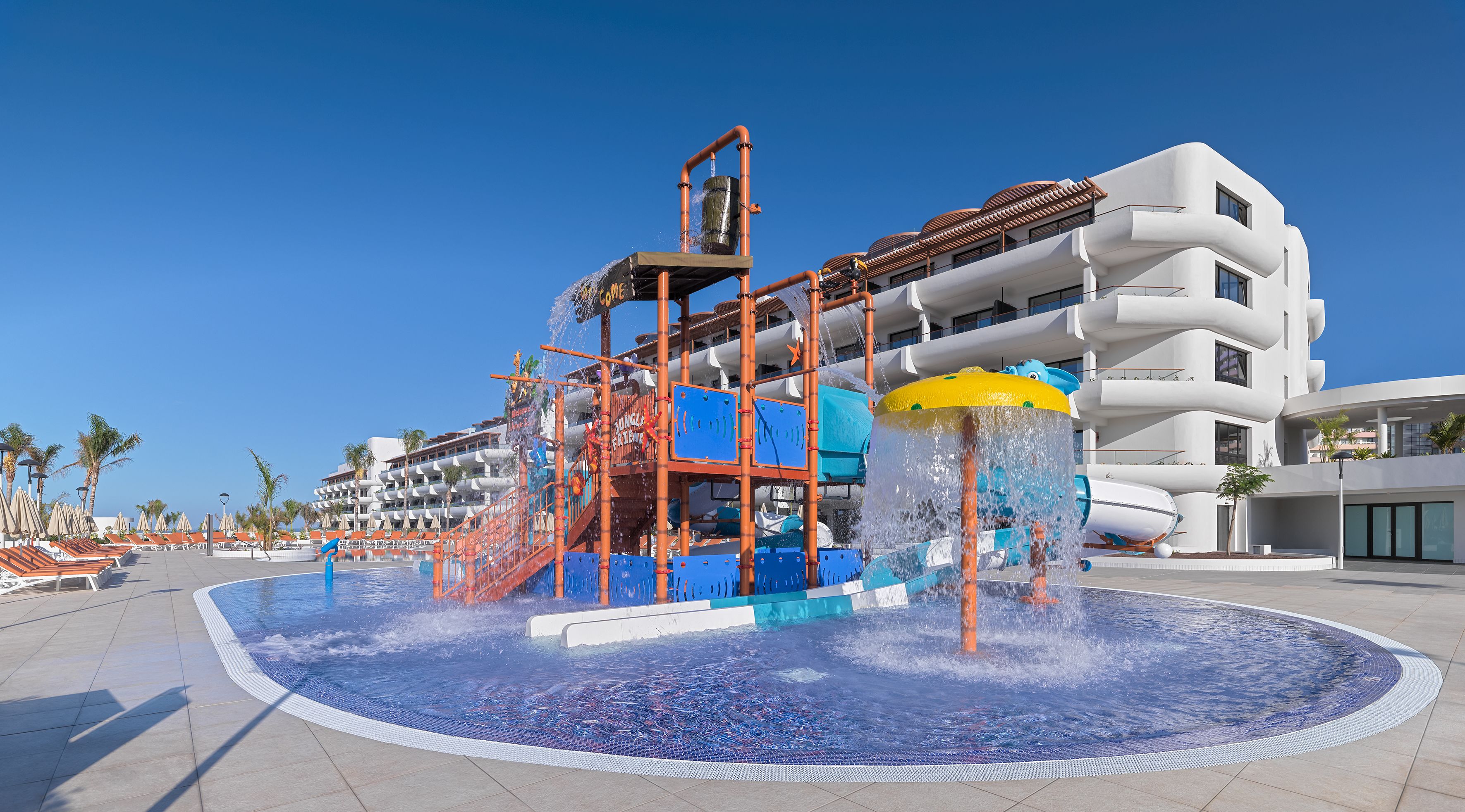Parque acuático con una gran fuente, una piscina y un edificio moderno con balcones.
