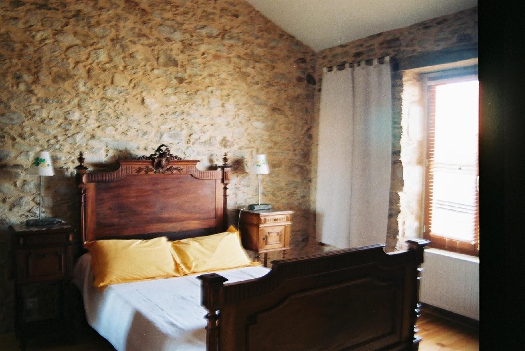 Een slaapkamer met een stenen muur, een houten bed met hoofdeinde, twee nachtkastjes met lampen en een raam met gordijnen.
