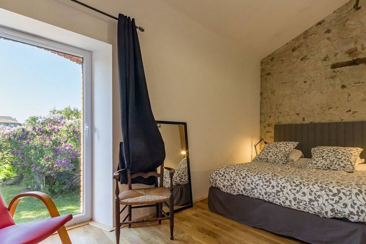 A bedroom with a large bed, a chair, a mirror, and a view of a garden through a sliding door.