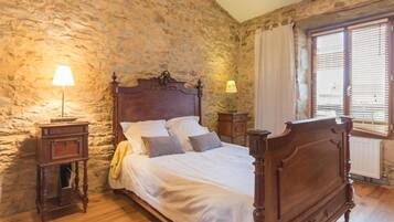 Une chambre avec un mur en pierre, un lit en bois, deux tables de chevet avec des lampes et une fenêtre avec des stores.