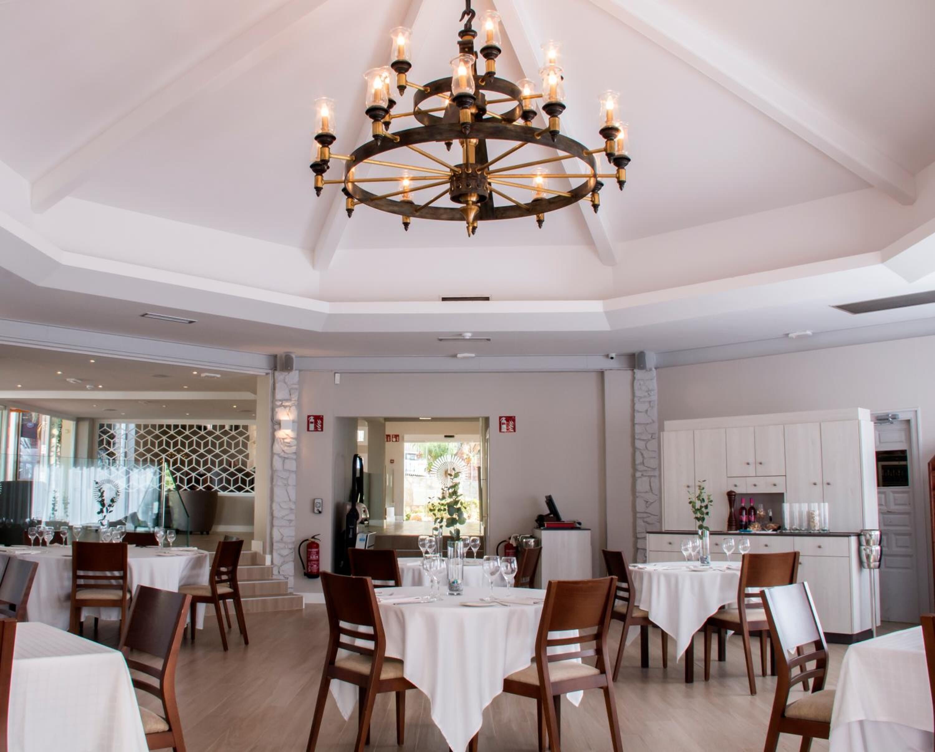 Un vaste espace dédié à la salle à manger, avec plusieurs tables et chaises, un lustre central et une vue sur la cuisine.