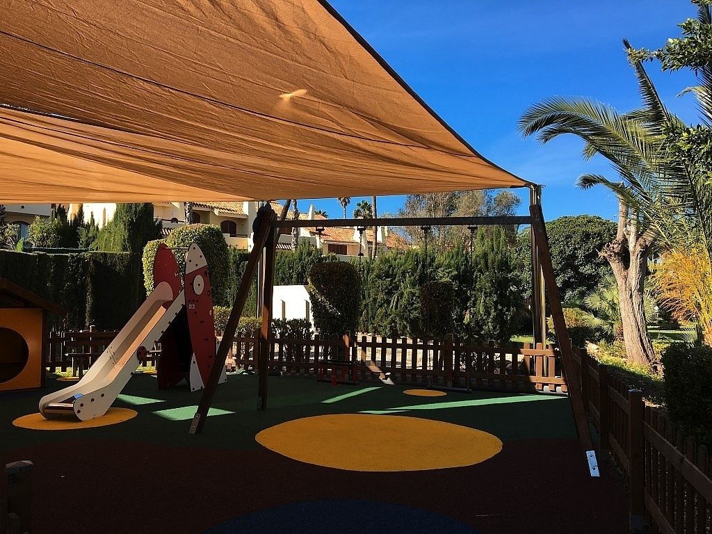 A playground with a slide and climbing structures, surrounded by a wooden fence and palm trees.