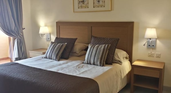 A neatly made bed with a wooden headboard, two bedside tables with lamps, and a window with curtains. - La Quinta at La Manga Club (Cartagena)