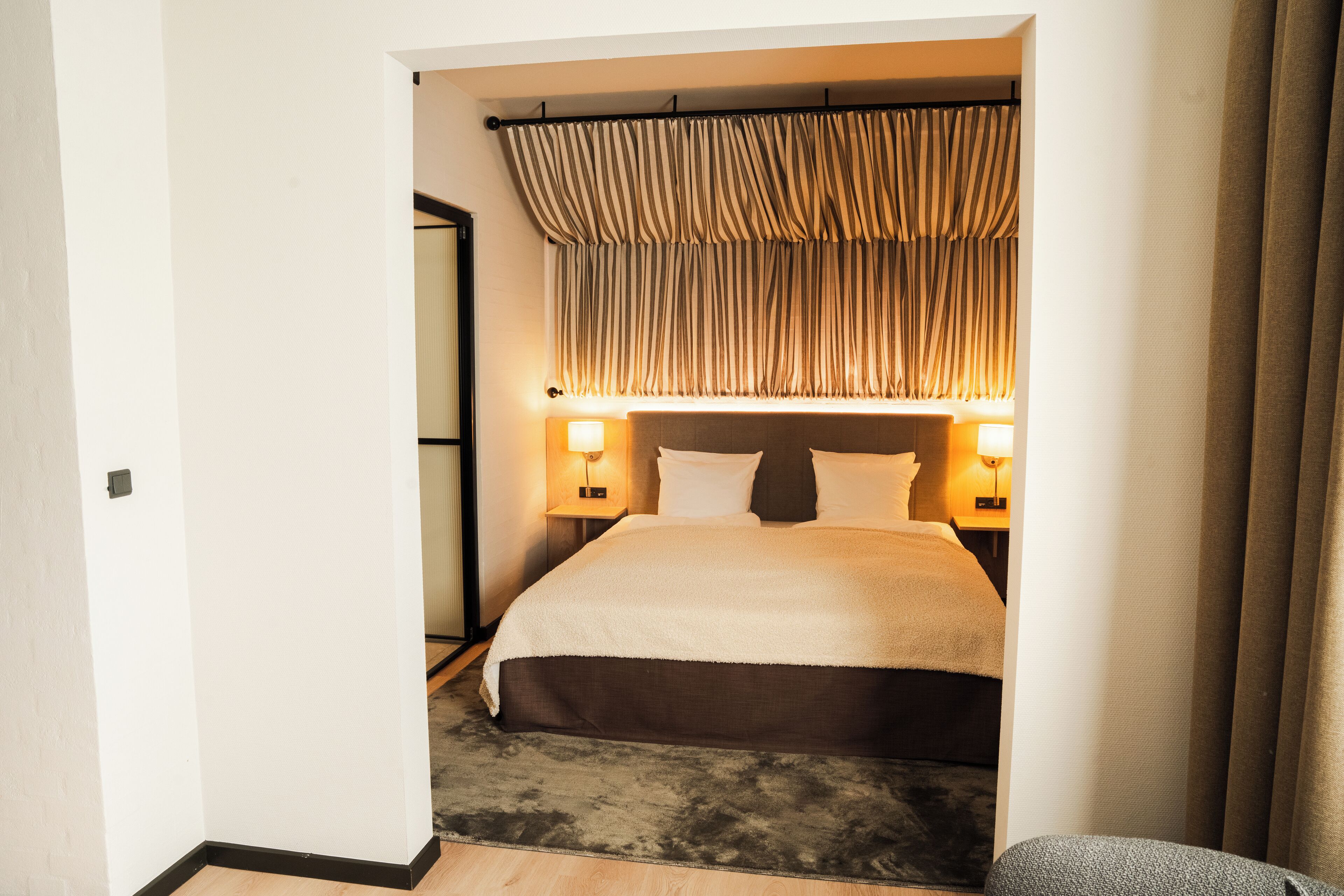 A hotel room with a large bed, two bedside lamps, a headboard with curtains, and a carpeted floor.