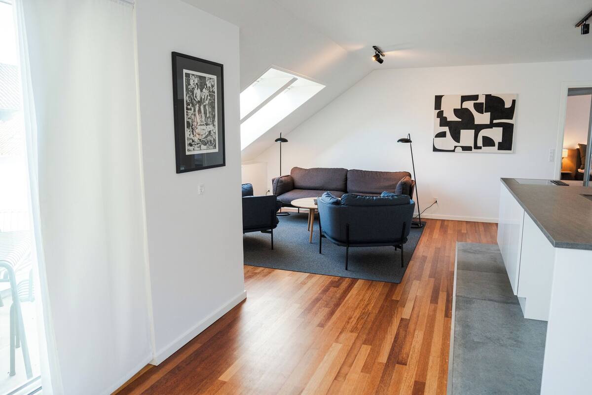 A modern living room with a wooden floor, a gray sofa, a small table, and a black floor lamp. There is a framed picture on the wall and a skylight above the sofa.