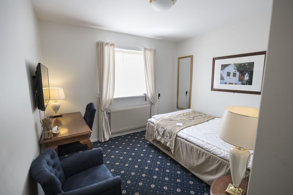 A hotel room with a bed, a desk, a chair, a lamp, a radiator, and a window with curtains.