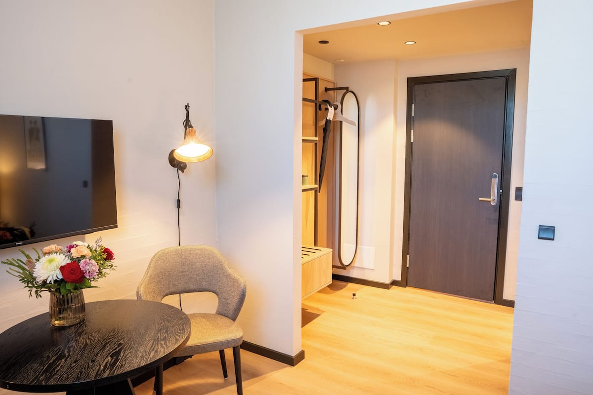 A modern hotel room with a dark wood floor, a round table with a vase of flowers, a grey upholstered chair, a wall-mounted TV, and a door leading to a shower area.