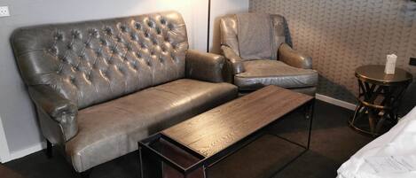 A modern hotel room with a leather sofa, a matching armchair, a wooden coffee table, and a small round table with a lamp.