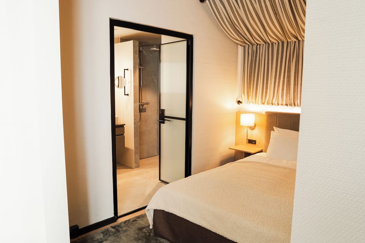 A hotel room with a bed, a nightstand with a lamp, a door leading to a bathroom, and a striped curtain.