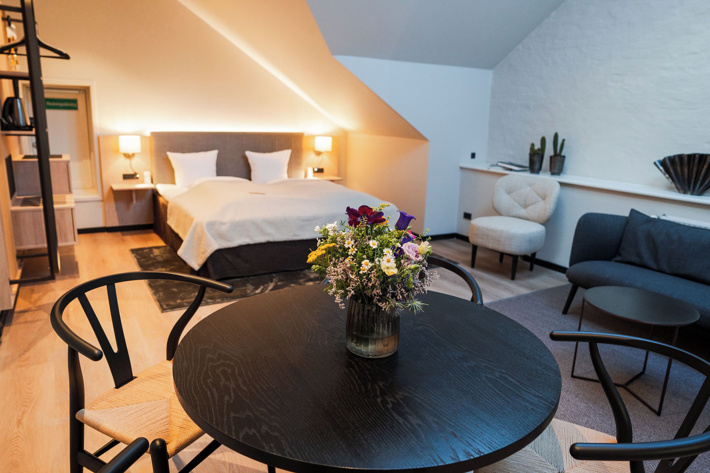 A modern hotel room with a bed, bedside tables, a small table with a vase of flowers, a sofa, and a chair.