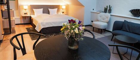 A modern hotel room with a bed, bedside tables, a small table with a vase of flowers, a sofa, and a chair.