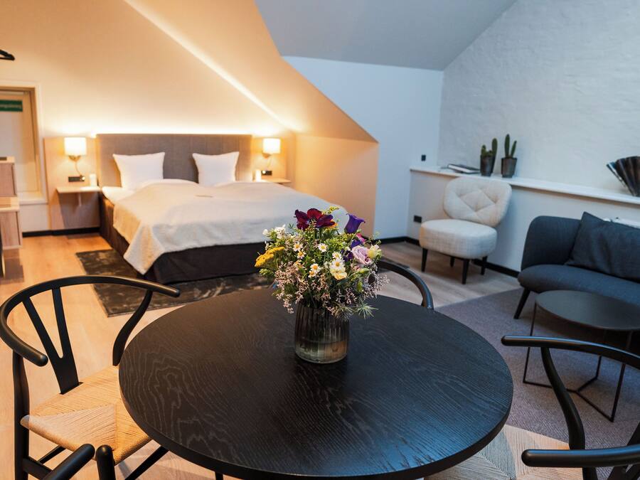 A modern hotel room with a bed, bedside tables, a small table with a vase of flowers, a sofa, and a chair.
