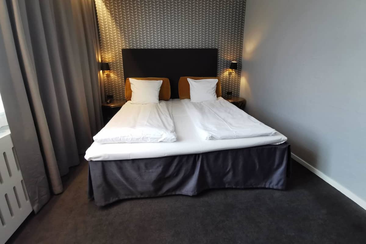 A neatly made bed with white linens, a dark skirted bed base, and a patterned wall behind it.