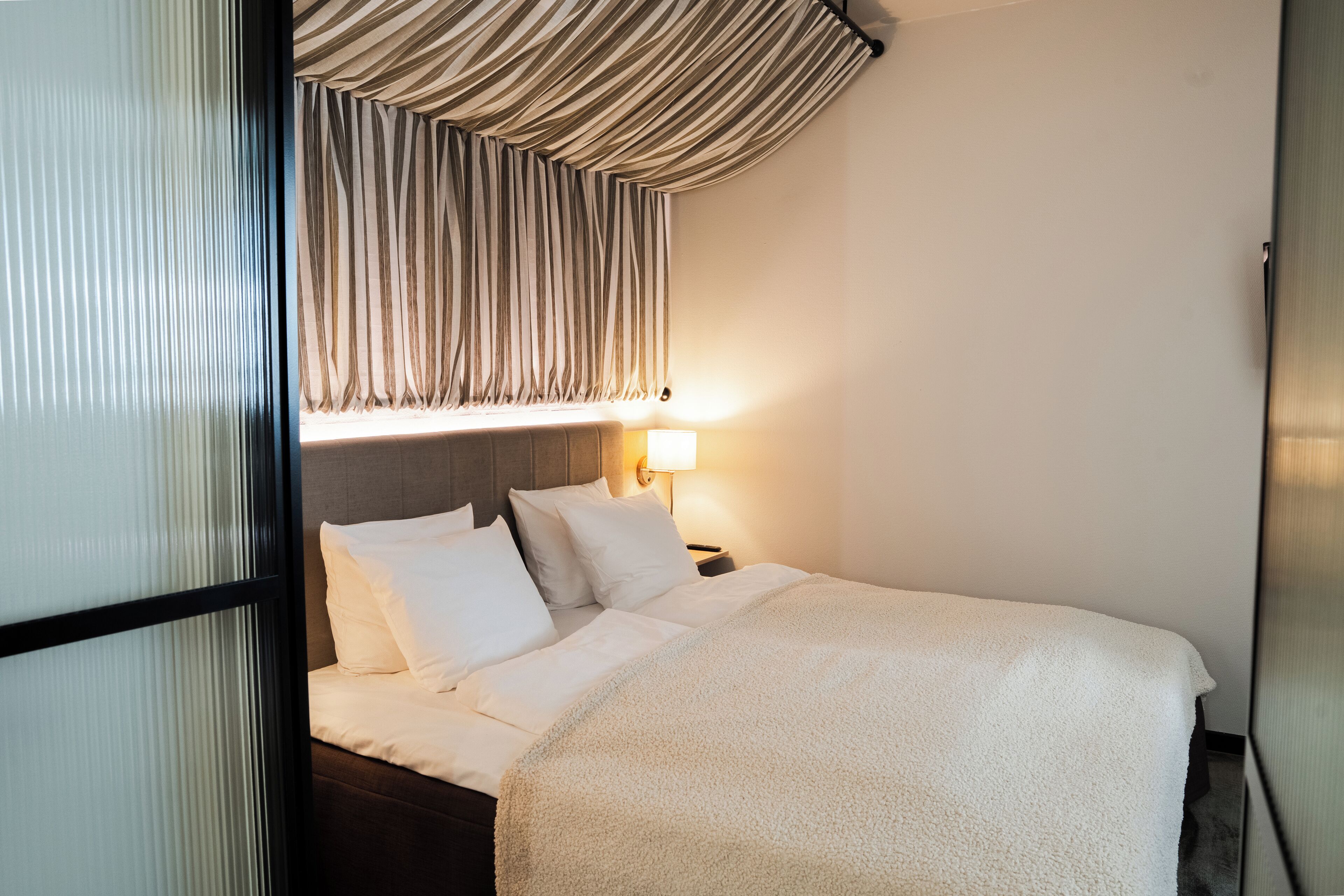 Una camera d'albergo con un letto, un comodino, una lampada e una finestra con tende.