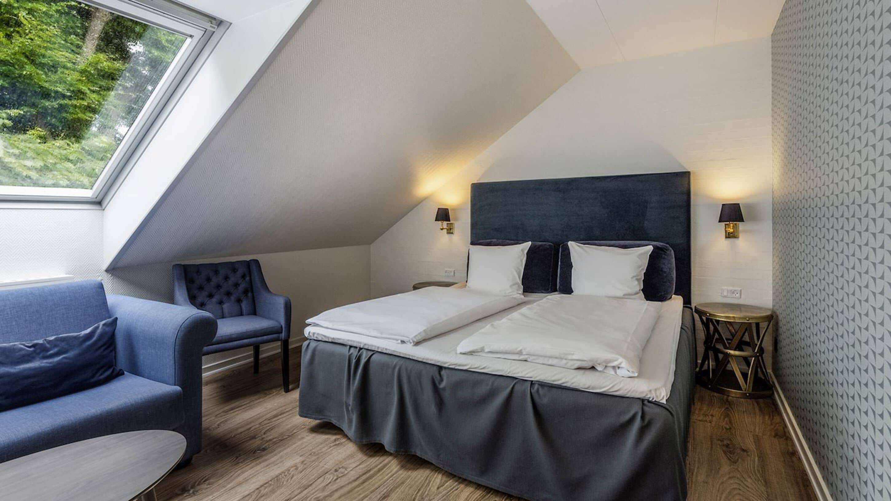 A modern bedroom with a sloped ceiling, a bed with white linens, a blue upholstered chair, and a small round table.