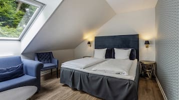 A modern bedroom with a sloped ceiling, a bed with white linens, a blue upholstered chair, and a small round table.