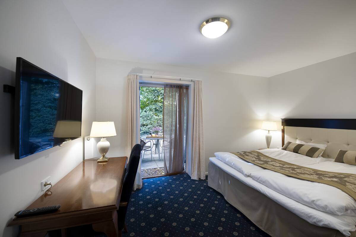 A hotel room with a bed, a desk with a lamp, a chair, a TV mounted on the wall, and a view of the outdoors through a door.