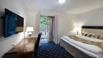 Habitación de hotel con cama, escritorio con lámpara, silla, televisor montado en la pared y vistas al exterior a través de una puerta.