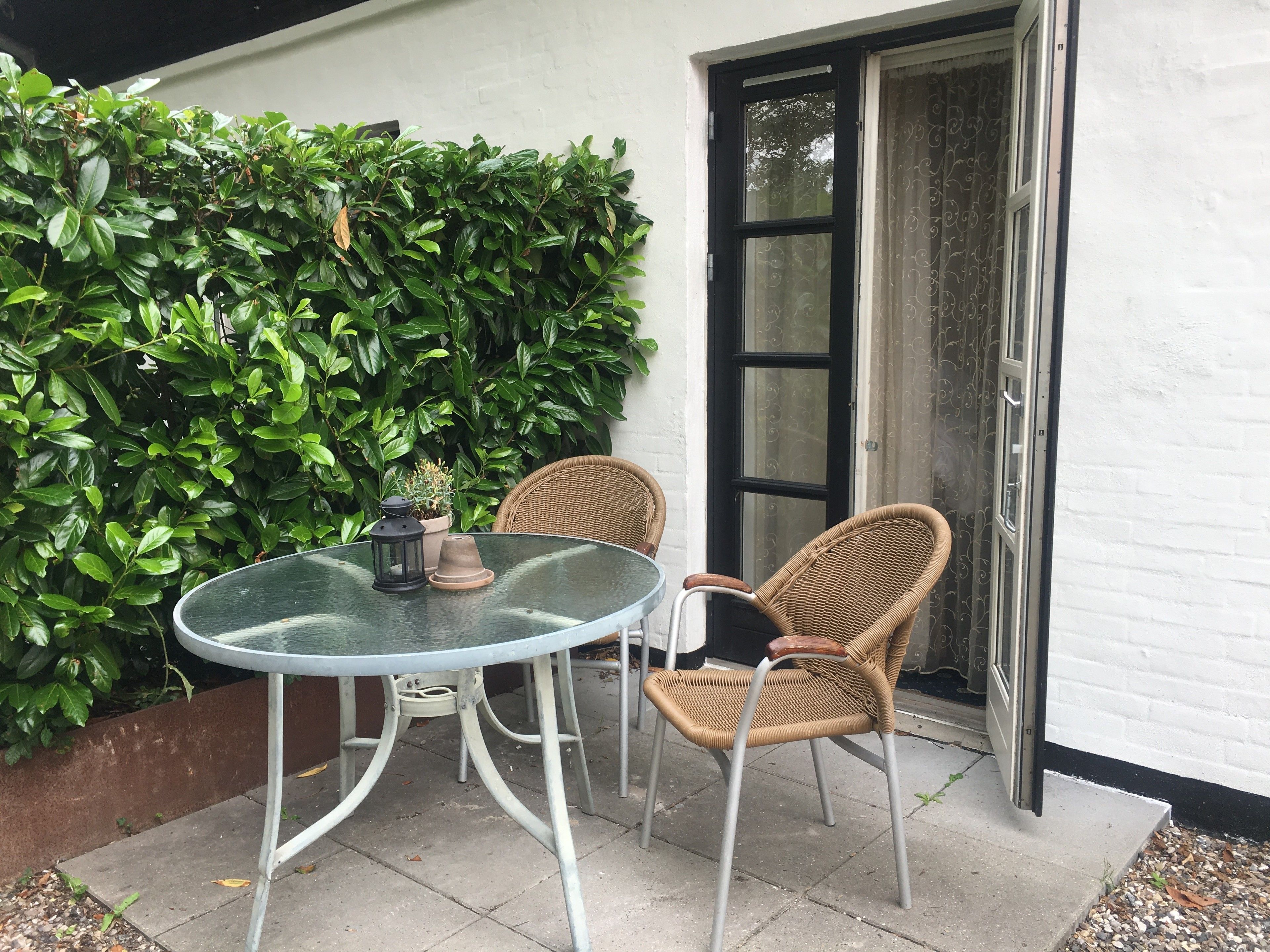 Een glazen tafel met twee rieten stoelen, een potplant en een deur met gordijn.