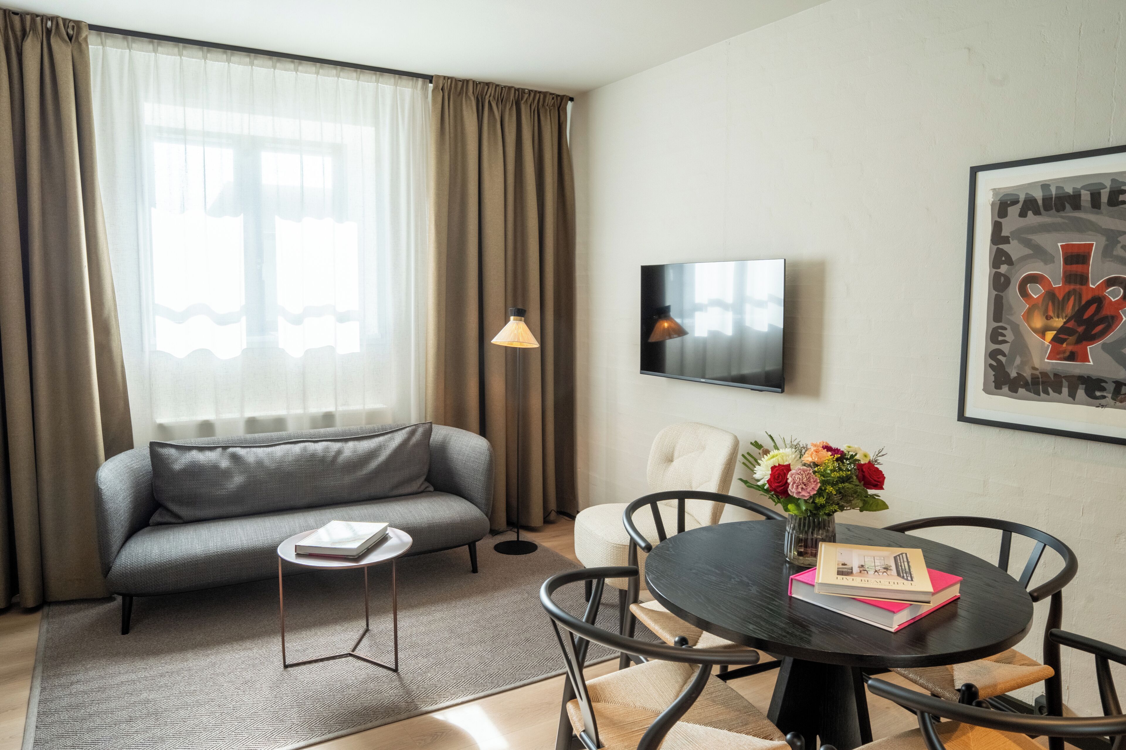 Una sala de estar moderna con un sofá gris, una mesa redonda de comedor con sillas, una pequeña mesita auxiliar, una lámpara de pie y una obra enmarcada en la pared.