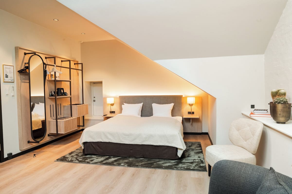 A bedroom with a bed, bedside tables, a chair, a mirror, and a shelf with books and a plant.