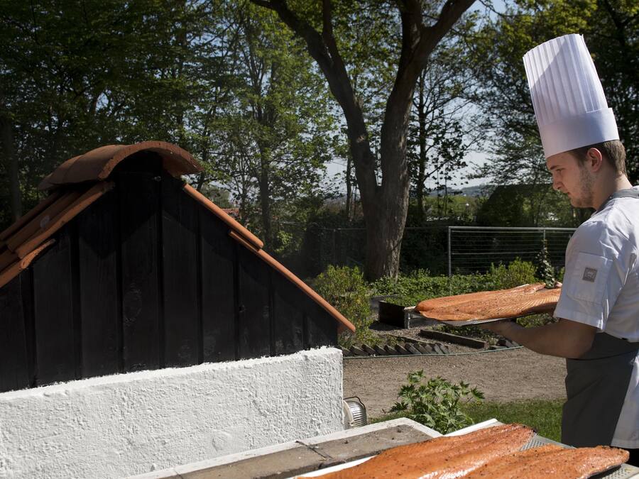 A chef holding a large piece of salmon on a grill outdoors, with a wooden structure and trees in the background.