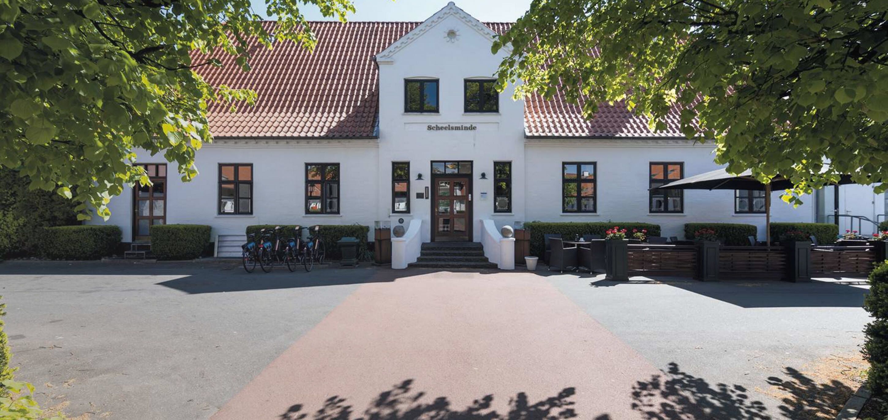 A white building with a red-tiled roof, named "Scheelsminde," featuring a central entrance, a small staircase, and a paved driveway.