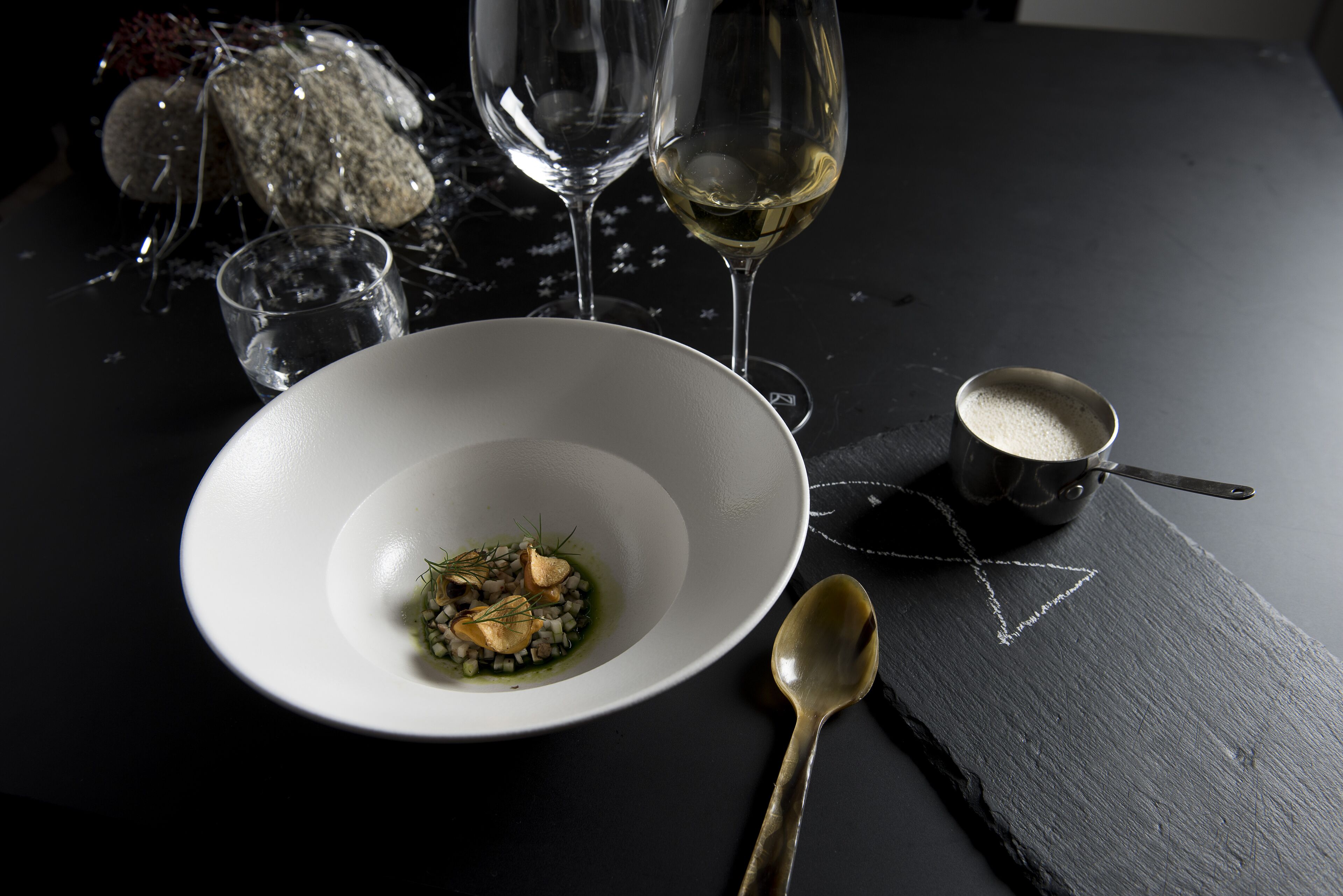 A white bowl with a spoon on a dark surface, accompanied by a small bowl and a glass of white wine.