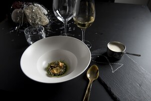 Un tazón blanco con una cuchara sobre una superficie oscura, junto a un tazón pequeño y una copa de vino blanco.