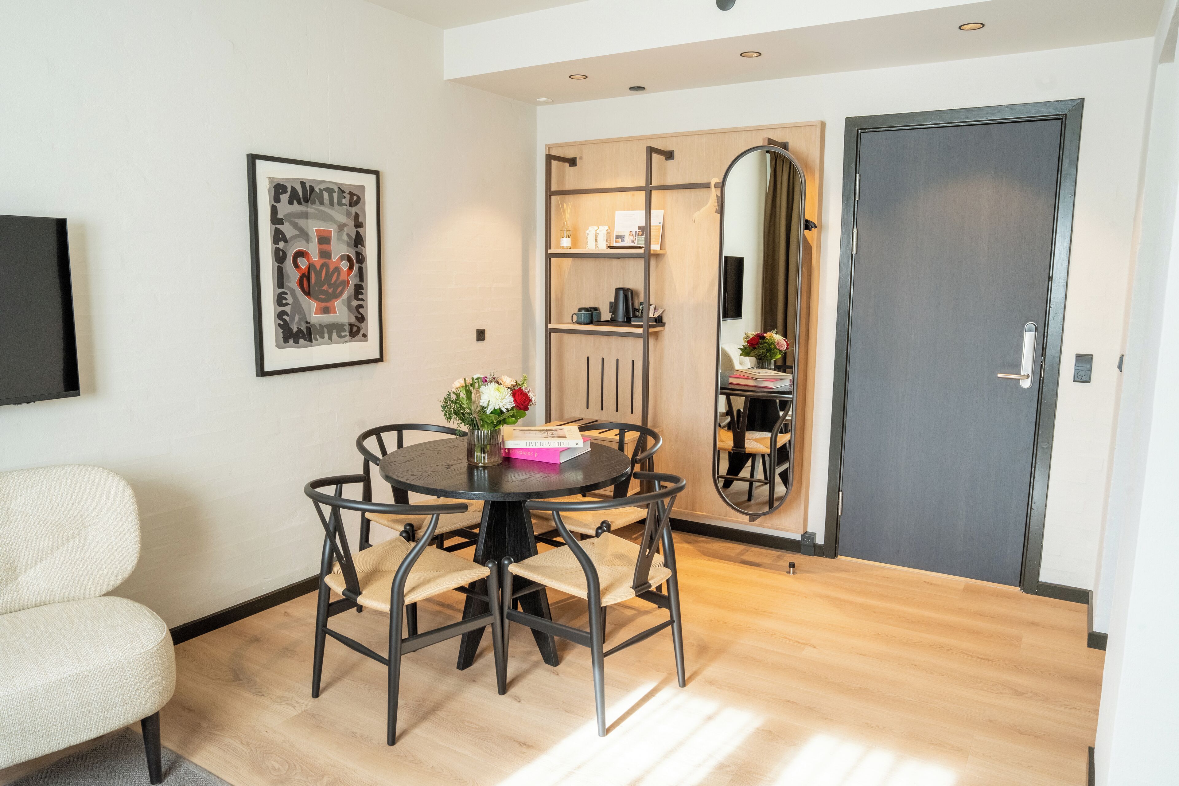 A dining area with a round table, four chairs, a vase with flowers, and a framed picture on the wall.