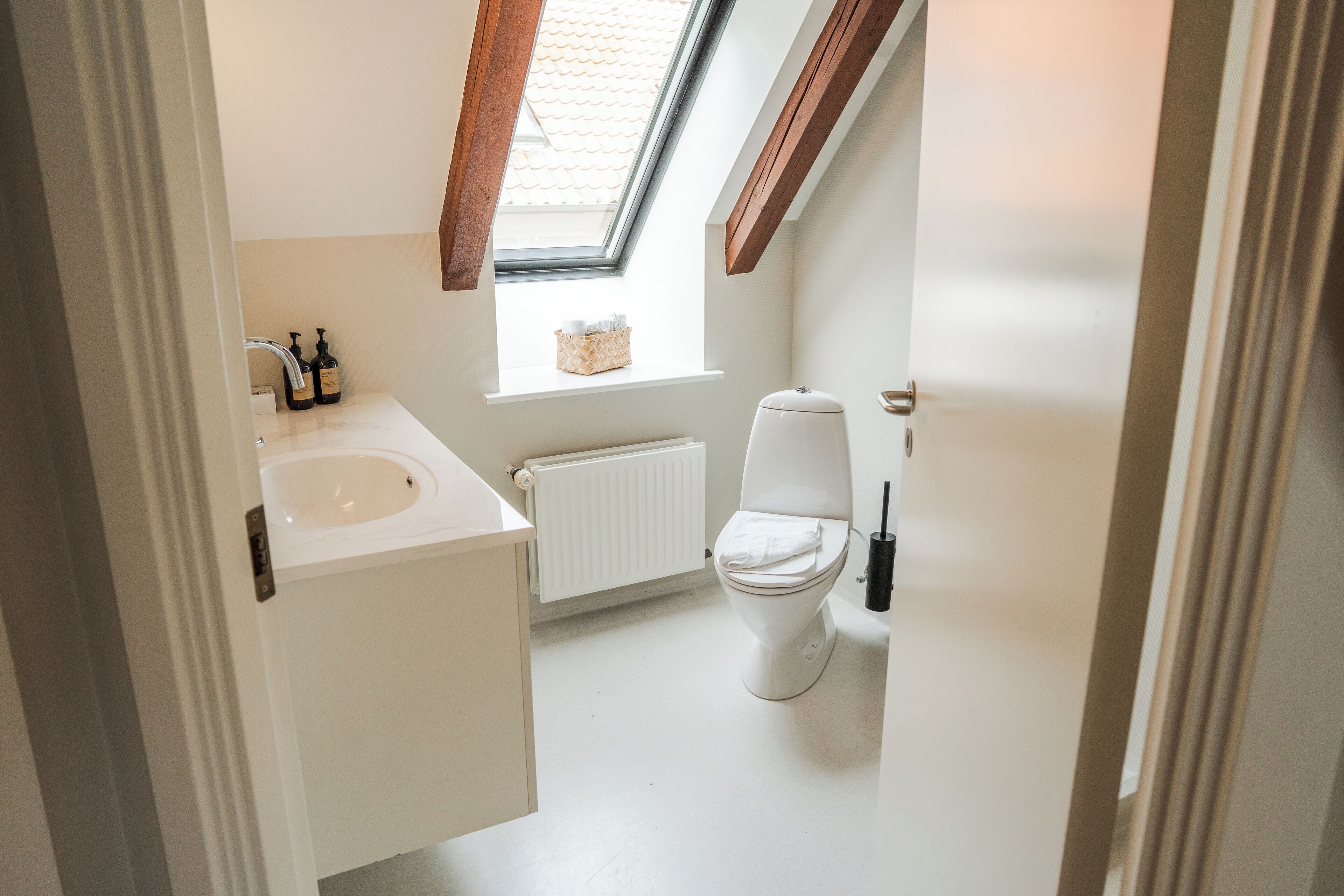 Une petite salle de bain lumineuse, dotée d’un puits de lumière, d’un lavabo blanc et de toilettes.