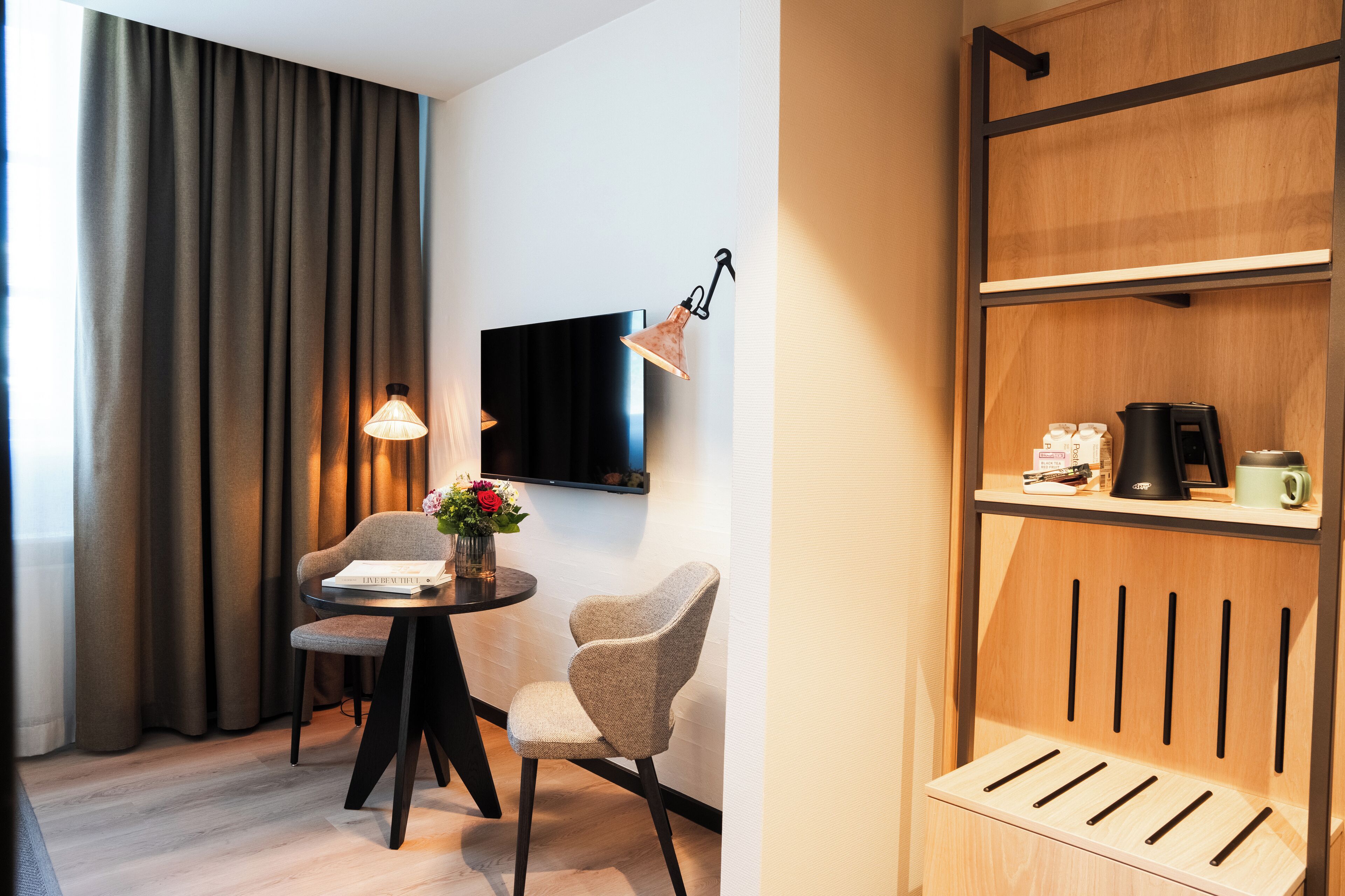 Et hotelværelse med et fladskærms-tv på væggen, et rundt bord med to stole, en vase med blomster samt en hylde med kaffemaskine og andet udstyr.