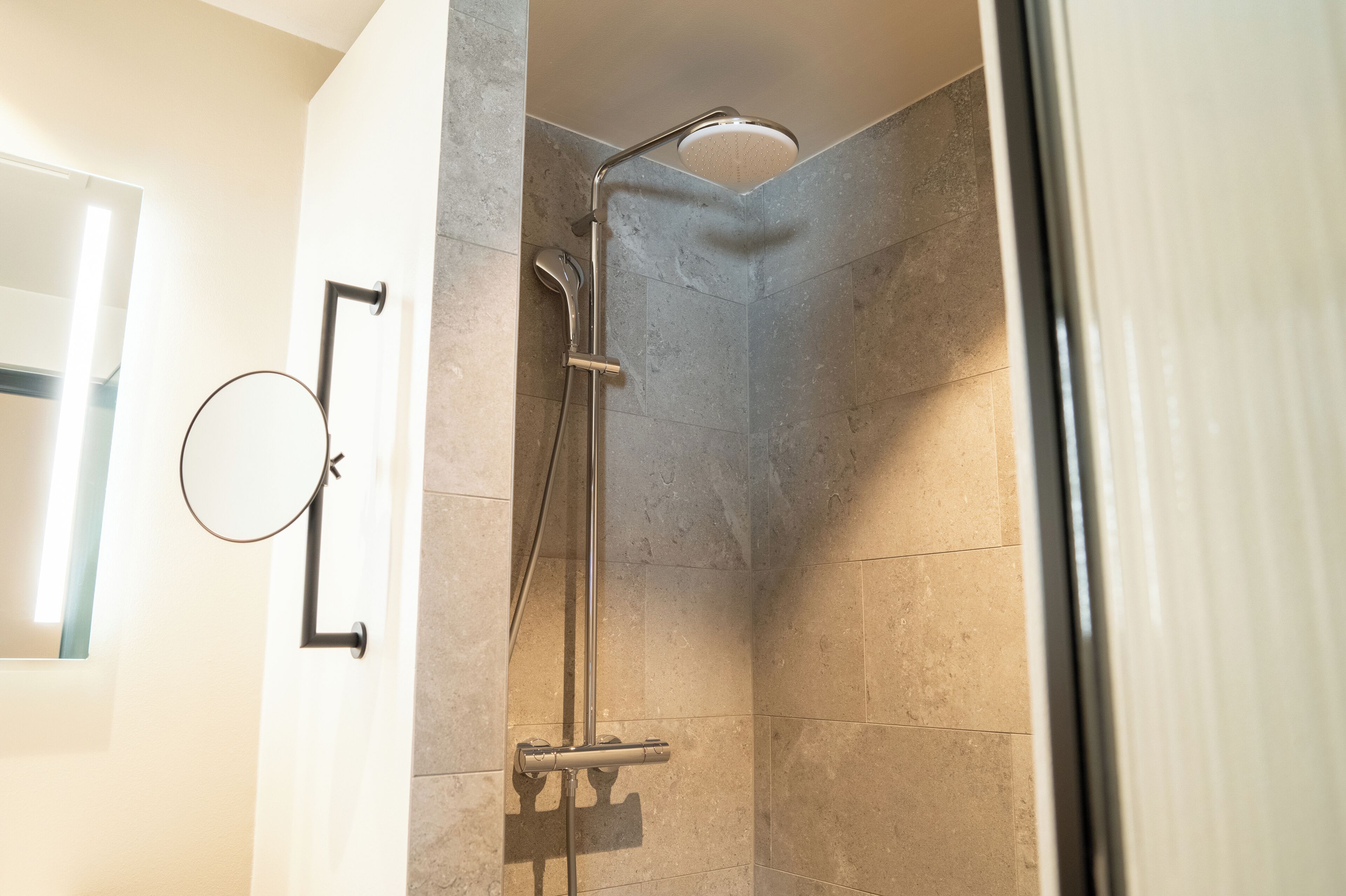 A shower with a handheld showerhead and a wall-mounted faucet in a corner of a room with tiled walls.